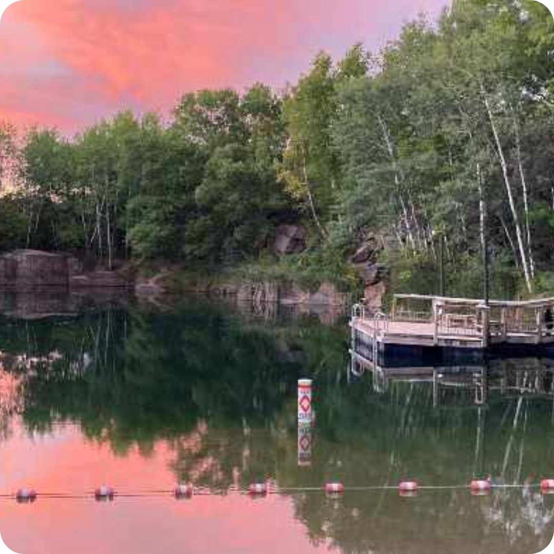 A quarry lake at twilight with a pink-orange sky, reflecting trees and a wooden dock on the right, alongside a warning buoy and red-and-white boundary buoys.